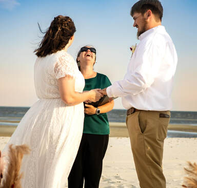 A wedding officiant in a green shirt and sunglasses stands between a bride and groom on a sandy beach, holding their hands during a ceremony.