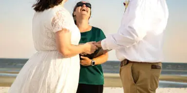A wedding officiant in a green shirt and sunglasses stands between a bride and groom on a sandy beach, holding their hands during a ceremony.