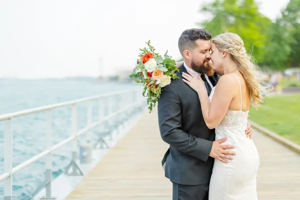 A bride and groom embracing on a wooden pier near a body of water, with the bride holding a bouquet of orange and white flowers.