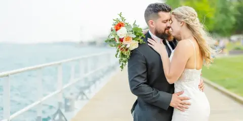 A bride and groom embracing on a wooden pier near a body of water, with the bride holding a bouquet of orange and white flowers.