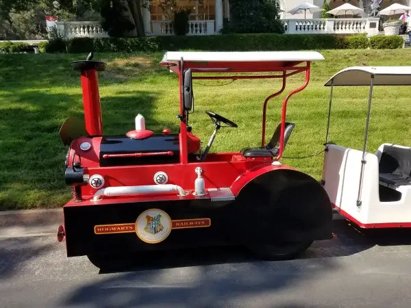 A red and black custom trackless train with a white canopy and passenger car parked on a paved surface.