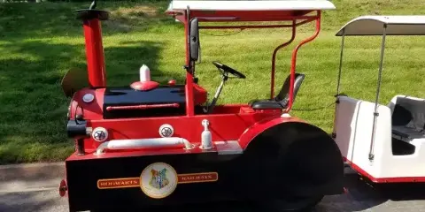 A red and black custom trackless train with a white canopy and passenger car parked on a paved surface.