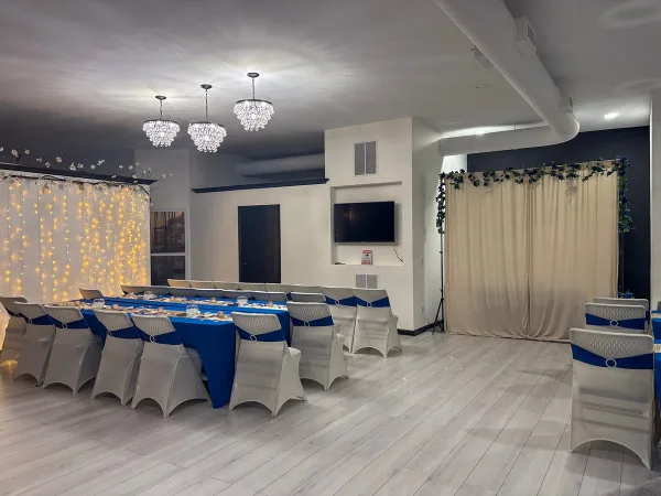 Interior view of an event venue with a long blue table, white chairs, and decorative lighting.