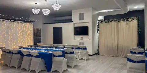 Interior view of an event venue with a long blue table, white chairs, and decorative lighting.