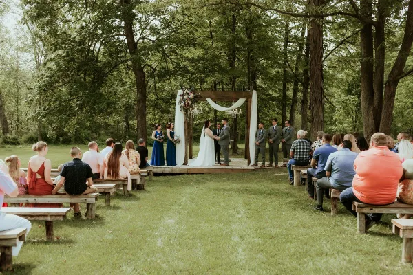 A wedding ceremony taking place outdoors in a wooded area with a wooden arch and guests seated on benches.