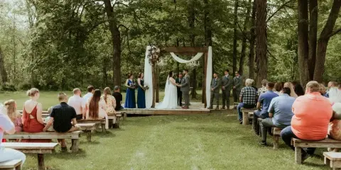 A wedding ceremony taking place outdoors in a wooded area with a wooden arch and guests seated on benches.