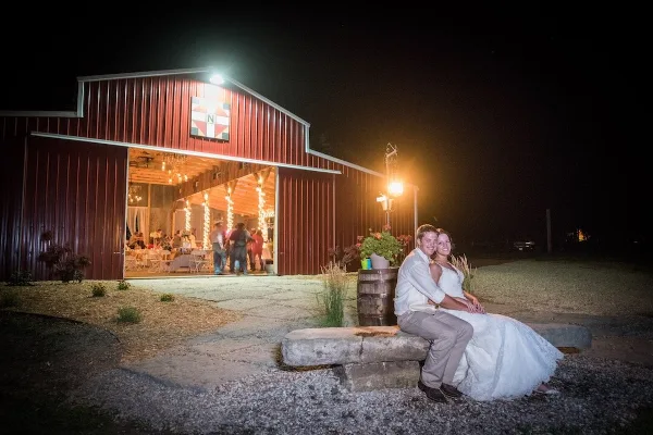 A red barn wedding venue with open doors showing a decorated interior and a couple posing outside at night.