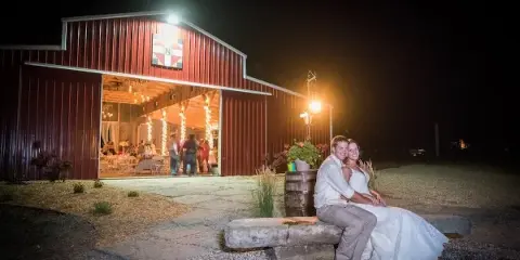 A red barn wedding venue with open doors showing a decorated interior and a couple posing outside at night.