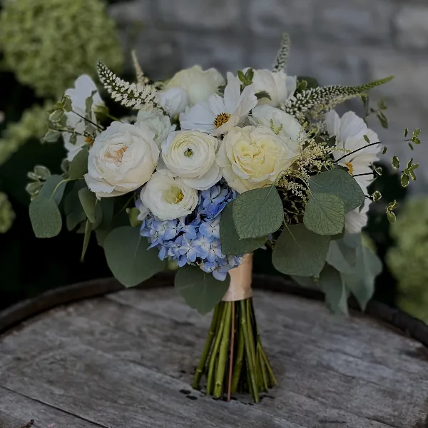 A wedding bouquet with white roses, ranunculus, and blue hydrangeas resting on a wooden barrel.