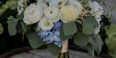 A wedding bouquet with white roses, ranunculus, and blue hydrangeas resting on a wooden barrel.