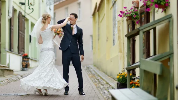 A bride and groom standing in a cobblestone street, with the bride holding a bouquet and the groom adjusting his glasses.