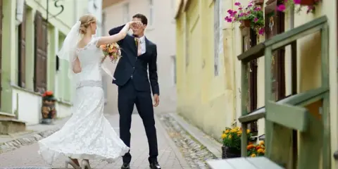 A bride and groom standing in a cobblestone street, with the bride holding a bouquet and the groom adjusting his glasses.