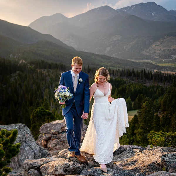 A bride and groom walking hand-in-hand on a rocky mountain overlook with a forested valley and mountain peaks in the background.