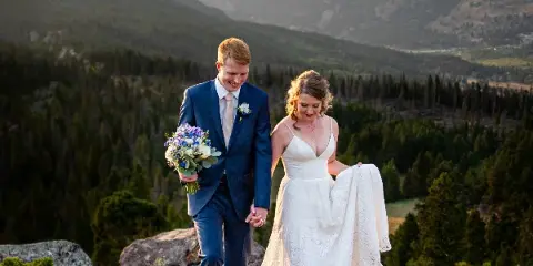 A bride and groom walking hand-in-hand on a rocky mountain overlook with a forested valley and mountain peaks in the background.