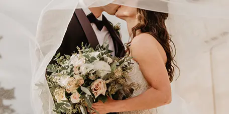 A bride and groom kissing under a veil, with the bride holding a large bouquet of white and peach flowers.