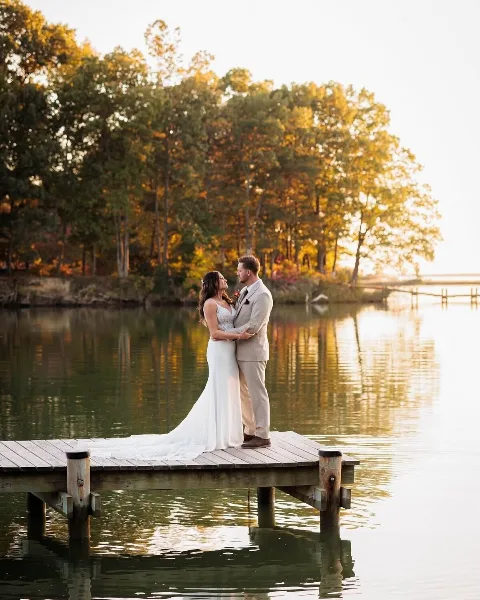 A bride and groom standing on a wooden dock by a lake at sunset.