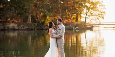 A bride and groom standing on a wooden dock by a lake at sunset.