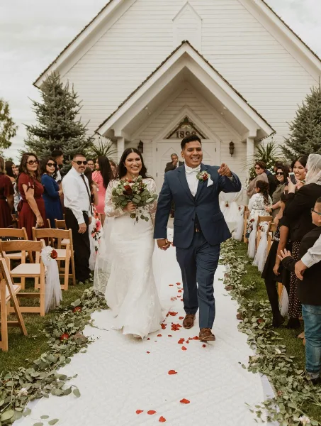 A bride and groom walking down the aisle at an outdoor wedding ceremony in front of a white chapel.