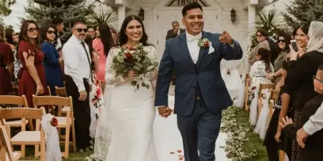 A bride and groom walking down the aisle at an outdoor wedding ceremony in front of a white chapel.