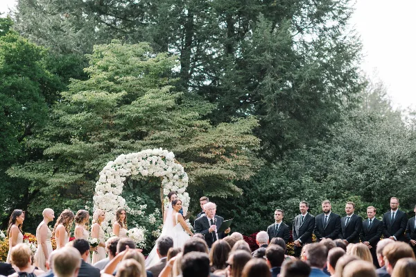 Outdoor wedding ceremony with bride and groom under a white floral arch, officiant speaking, and wedding party standing on either side.