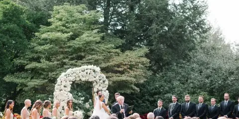 Outdoor wedding ceremony with bride and groom under a white floral arch, officiant speaking, and wedding party standing on either side.