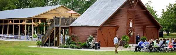A wide shot of a rustic red barn and wooden pavilion at a farm wedding venue with guests seated in chairs.