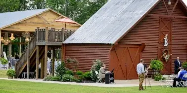 A wide shot of a rustic red barn and wooden pavilion at a farm wedding venue with guests seated in chairs.