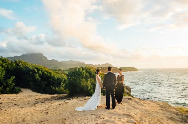 A bride and groom standing on a rocky coastal cliff facing an officiant during a wedding ceremony.