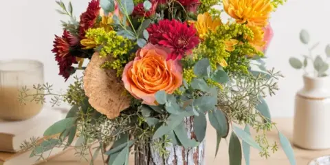 A mixed bouquet featuring orange roses, red dahlias, and greenery in a bark-covered vase.