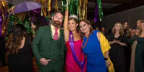 A professional photograph of a bride and groom at a festive event, surrounded by guests and colorful streamers.