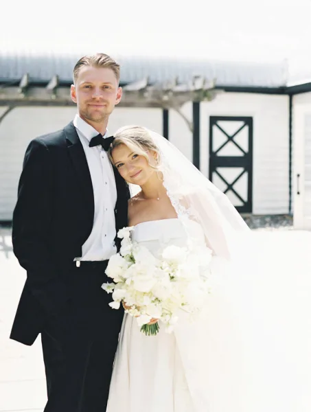 Bride and groom posing together outdoors with a white bouquet in front of a white barn-style building.