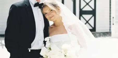 Bride and groom posing together outdoors with a white bouquet in front of a white barn-style building.