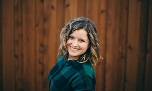 Portrait of Lacy Stone, a photographer, smiling in front of a wooden fence.