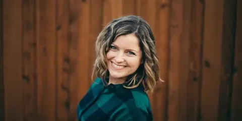 Portrait of Lacy Stone, a photographer, smiling in front of a wooden fence.