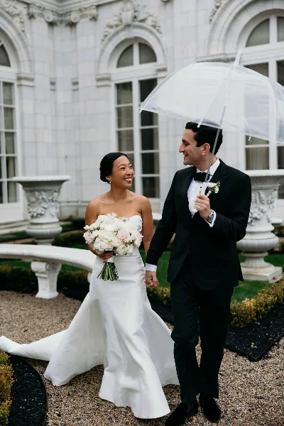 A bride in a white gown and a groom in a black tuxedo walking hand-in-hand under a clear umbrella in front of a white stone building.