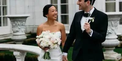 A bride in a white gown and a groom in a black tuxedo walking hand-in-hand under a clear umbrella in front of a white stone building.
