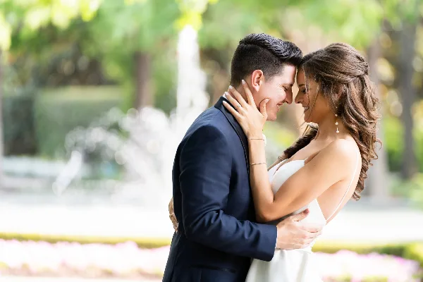 A bride and groom embracing in a garden setting with a fountain in the background.