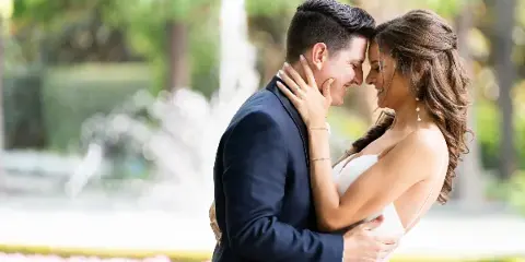 A bride and groom embracing in a garden setting with a fountain in the background.