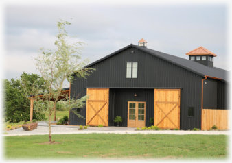 Exterior view of a large dark metal barn with wooden doors and a cupola, situated on a grassy lawn.