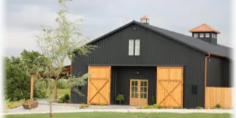 Exterior view of a large dark metal barn with wooden doors and a cupola, situated on a grassy lawn.