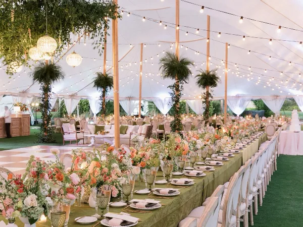 Luxury outdoor wedding reception inside a white tent featuring long tables with floral centerpieces, string lighting, chandeliers, and lounge furniture.