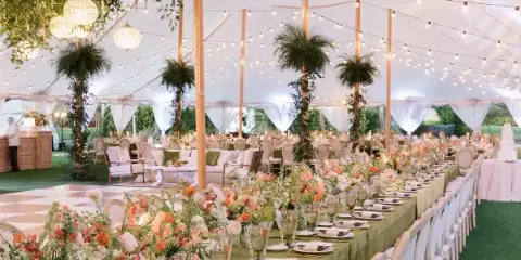 Luxury outdoor wedding reception inside a white tent featuring long tables with floral centerpieces, string lighting, chandeliers, and lounge furniture.