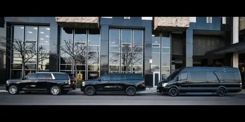 A lineup of black luxury SUVs and a passenger van parked in front of a modern glass building with the company name visible.