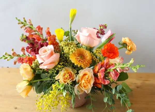 A colorful, handcrafted floral arrangement featuring roses, snapdragons, and other flowers in a vase on a wooden table.