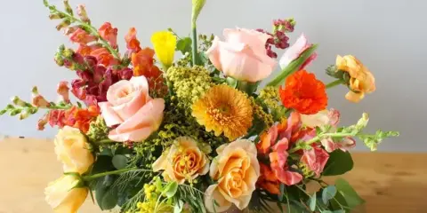 A colorful, handcrafted floral arrangement featuring roses, snapdragons, and other flowers in a vase on a wooden table.