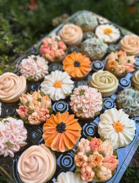 A close-up photograph of a tray filled with various decorated cupcakes featuring floral and rose designs in pastel and vibrant colors.