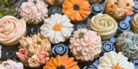 A close-up photograph of a tray filled with various decorated cupcakes featuring floral and rose designs in pastel and vibrant colors.