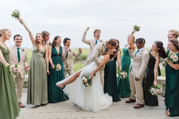 A bride and groom kissing on a wooden deck surrounded by their wedding party, with a coastal marsh landscape in the background.