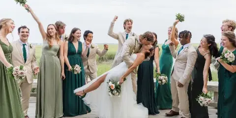 A bride and groom kissing on a wooden deck surrounded by their wedding party, with a coastal marsh landscape in the background.