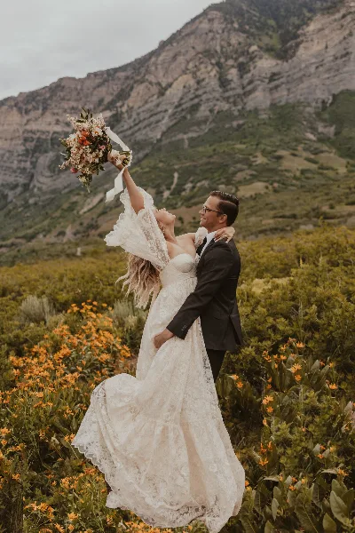 A bride and groom in a desert landscape with a mountain backdrop, the bride holding a bouquet aloft.